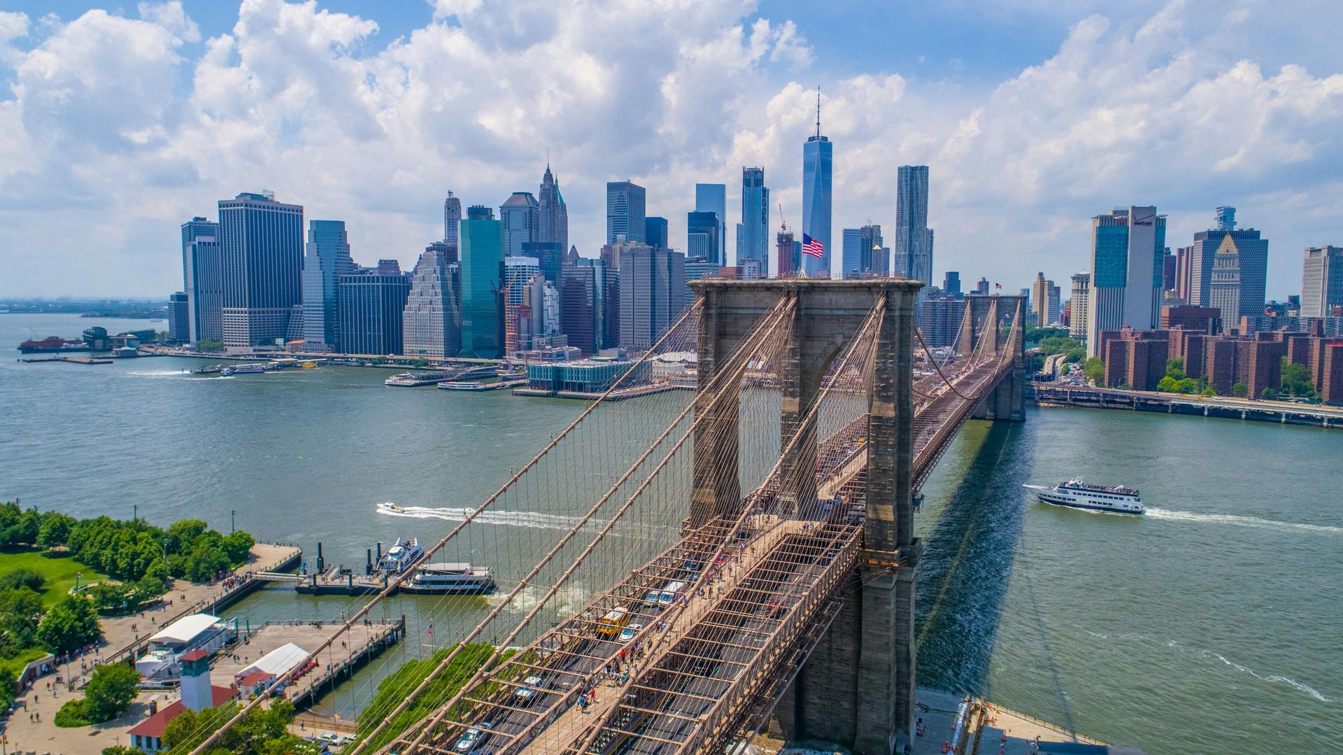 Brooklyn Bridge over water, cityscape in background, cloudy sky.