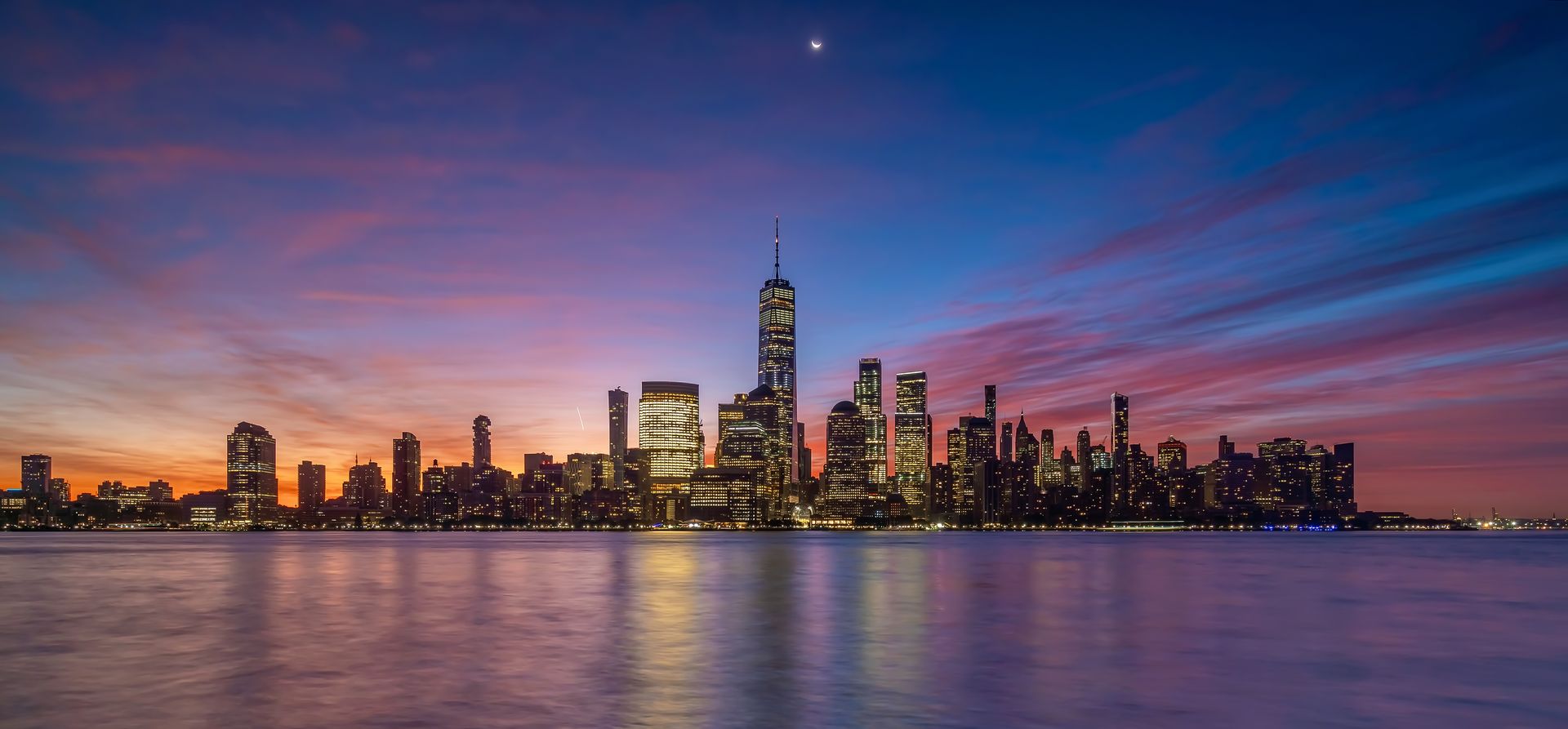 Skyline of New York City at sunset with buildings reflecting in the water below a colorful sky.