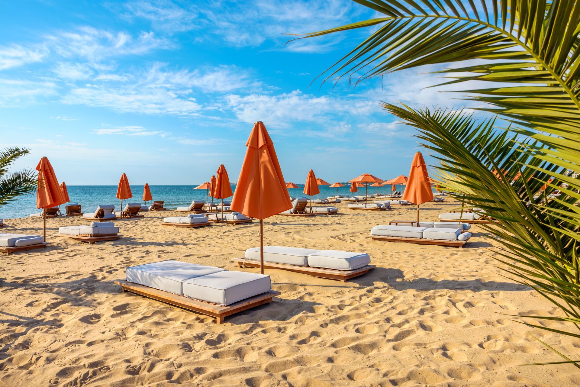 Beach scene with orange umbrellas, white lounge chairs on sand, blue sky.