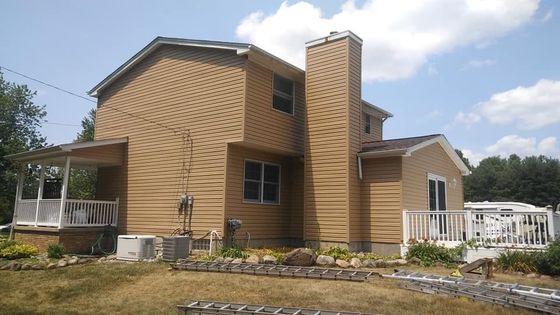 Two-story house with tan siding, chimney, porch, and white deck on a sunny day.