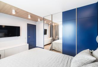 Una camera da letto moderna con pareti bianche, un armadio blu, un televisore e un guardaroba a specchio che riflette il letto.