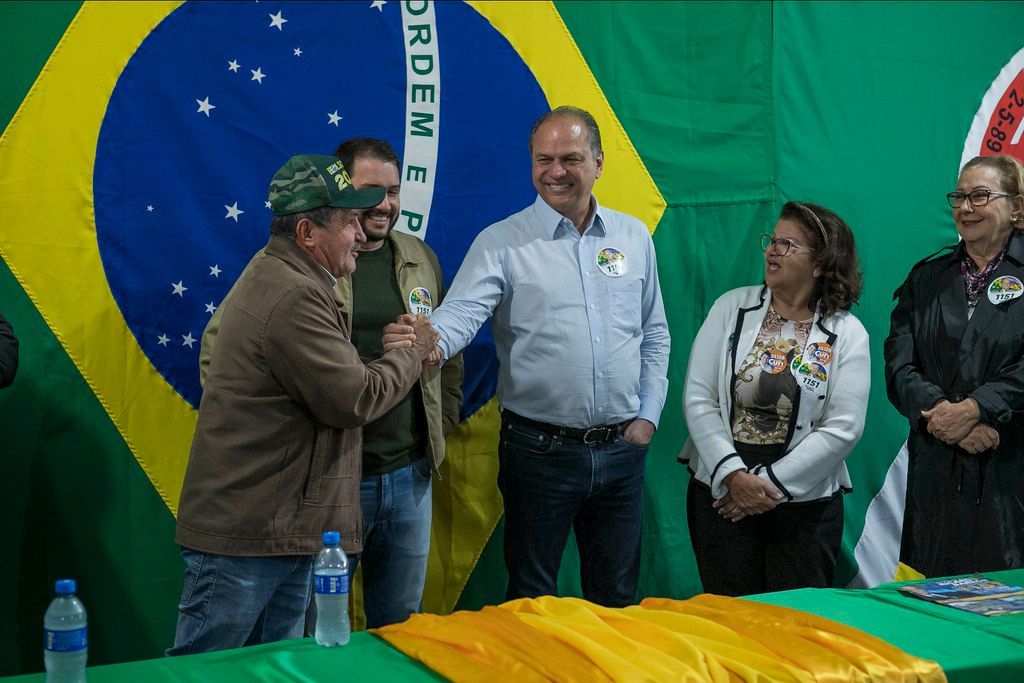 Um grupo de pessoas está em pé ao redor de uma mesa em frente a uma bandeira brasileira.