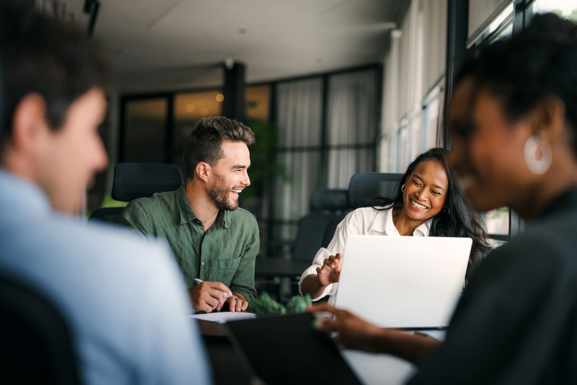 Colleagues smile and interact while collaborating at a desk with a laptop in a modern office.