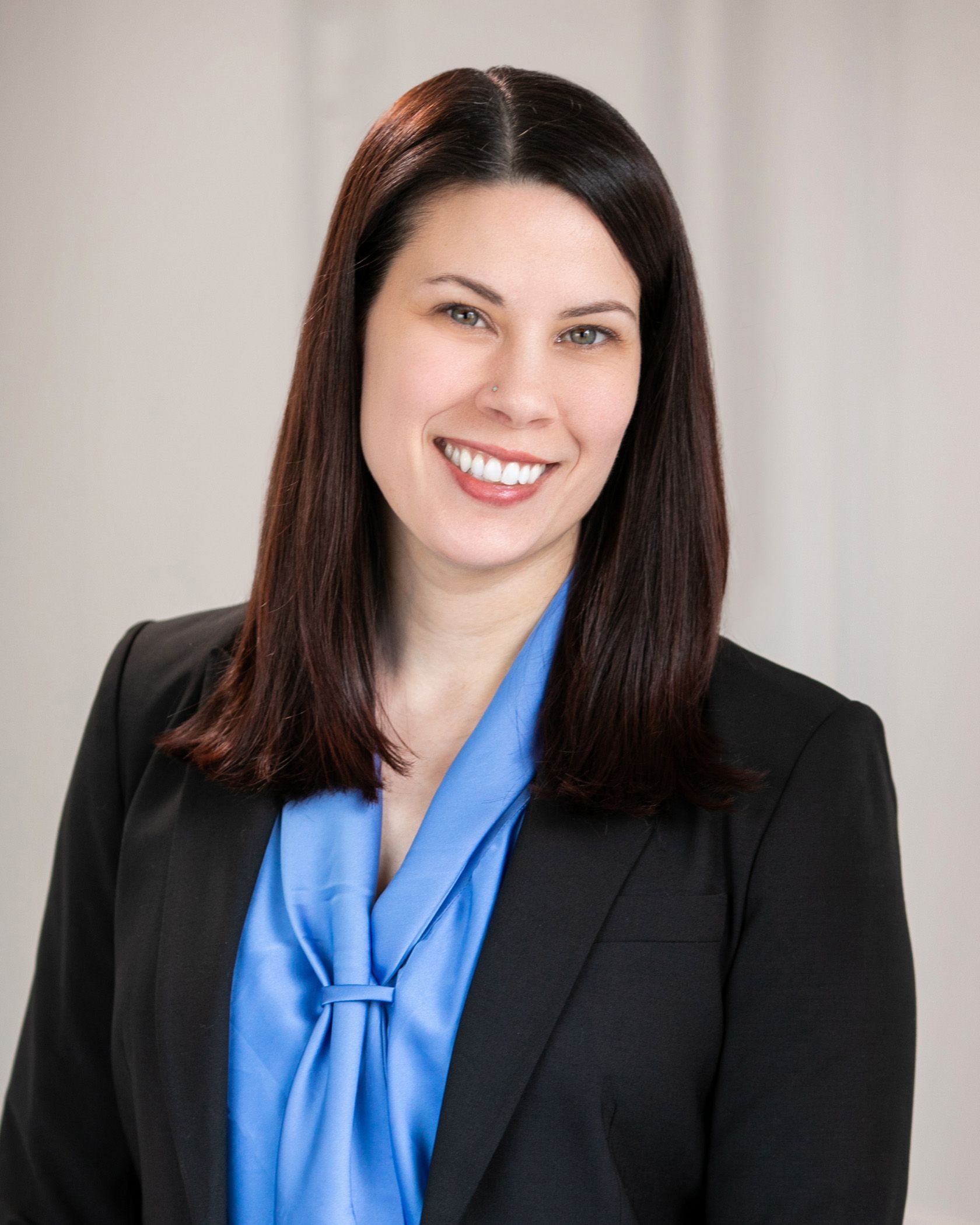 Woman with dark hair smiles, wearing a blue blouse and black blazer, against a light background.