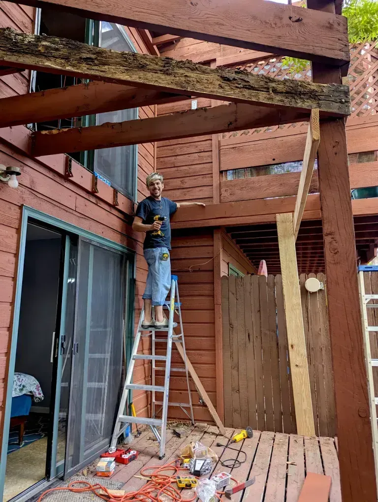 A man on a ladder drills into wood beams of an outdoor structure. He's wearing a blue shirt and looking at the camera.