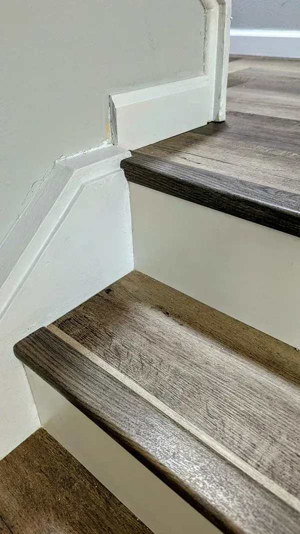 Staircase with wood-look treads, dark brown edges, and a light strip. White risers and wall, a corner is visible.