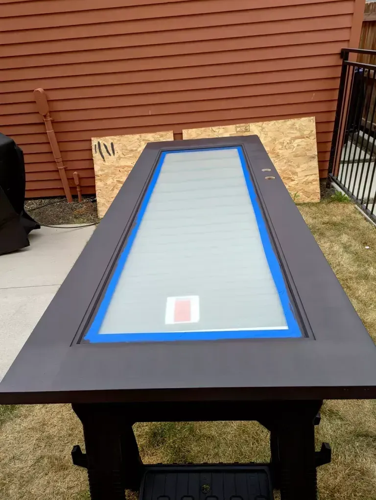 A door painted dark gray, taped around the glass panel, rests on a black sawhorse outdoors.