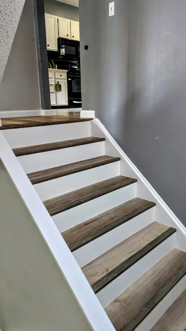 Staircase with white risers and wood-look treads leading towards a kitchen. Gray wall on the right.