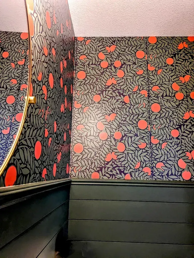 Wallpapered bathroom corner with black and coral floral pattern above dark green wainscoting. A round mirror is partially visible.