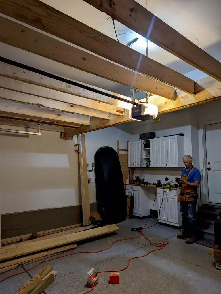 Man in garage with new ceiling beams. He's holding a phone. Boards are on the floor; garage door opener visible.
