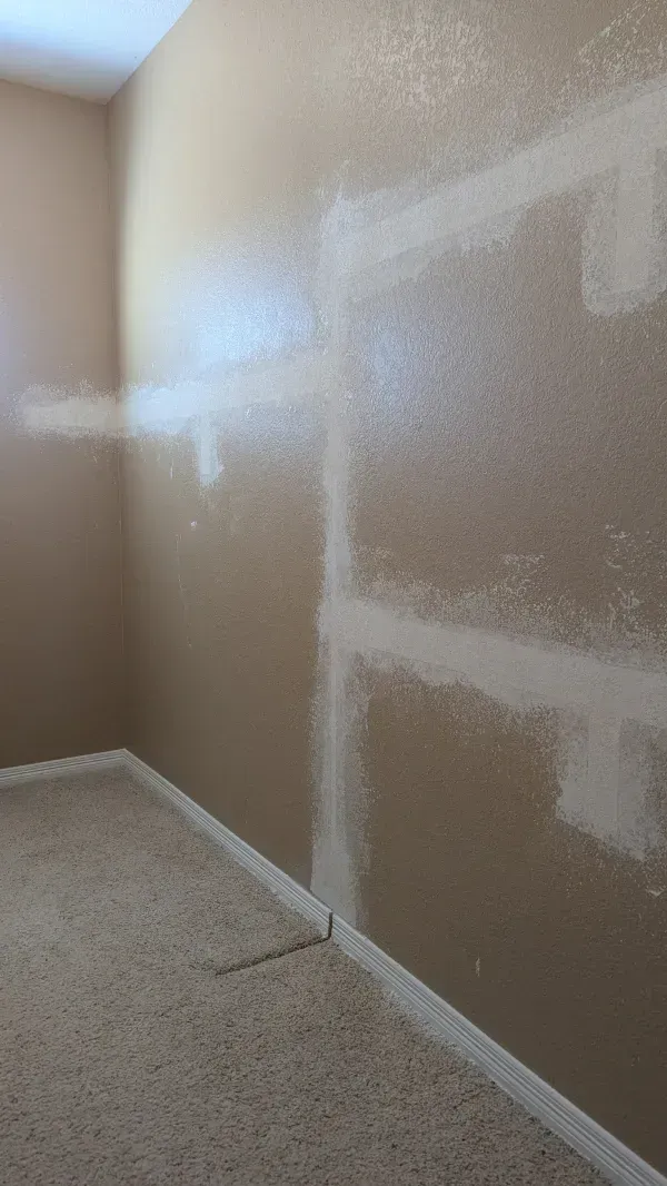 Beige interior corner, with patches of white plaster on the walls against a tan background, near a carpeted floor.