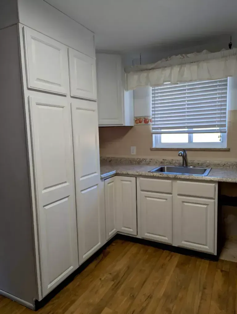 White kitchen cabinets and a sink in a room with a window, countertop, and wooden floor.