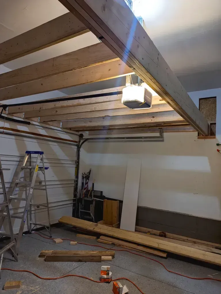 Garage under construction with exposed wooden beams overhead. A garage door, ladder, and materials are visible.