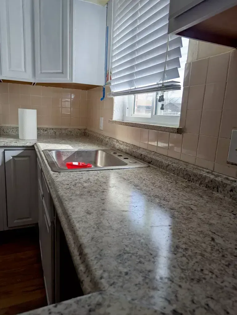 Kitchen with light-colored granite countertops, a stainless steel sink, white cabinets, and a window with closed blinds.