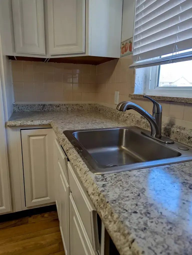 Kitchen sink with speckled countertops, white cabinets, and a window with blinds.