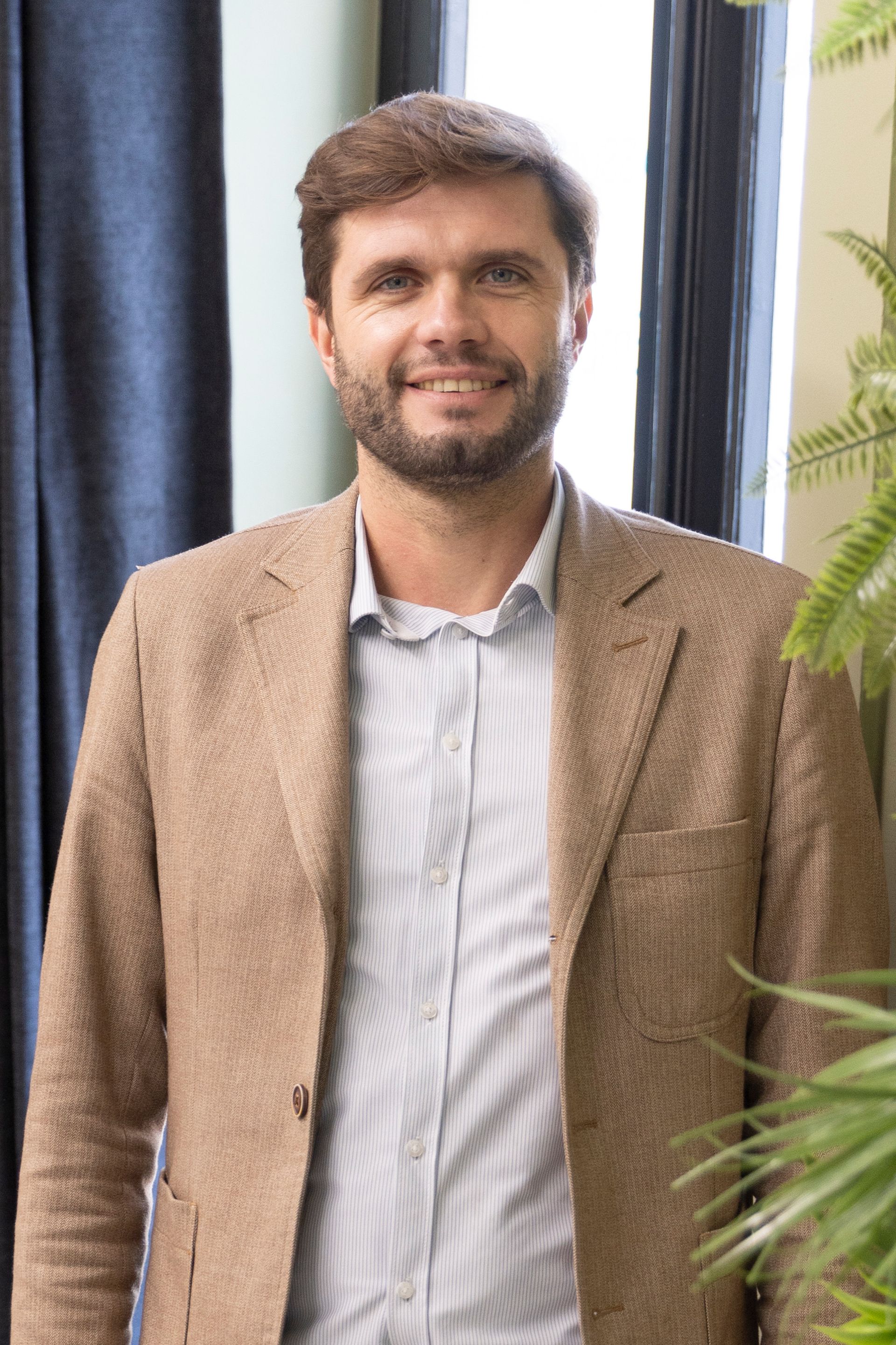 Un homme en blazer beige sourit, debout près d'une fenêtre avec une plante verte.