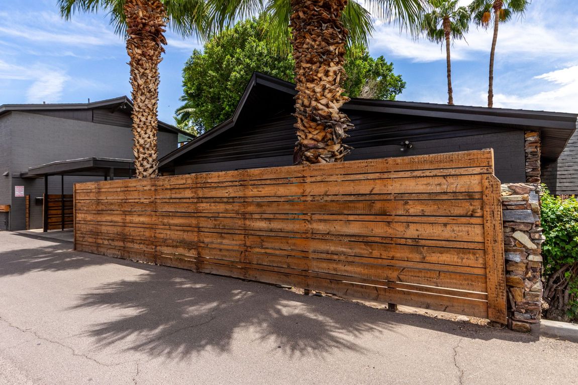 Wooden fence with palm trees and a dark-colored building behind it on a sunny day.