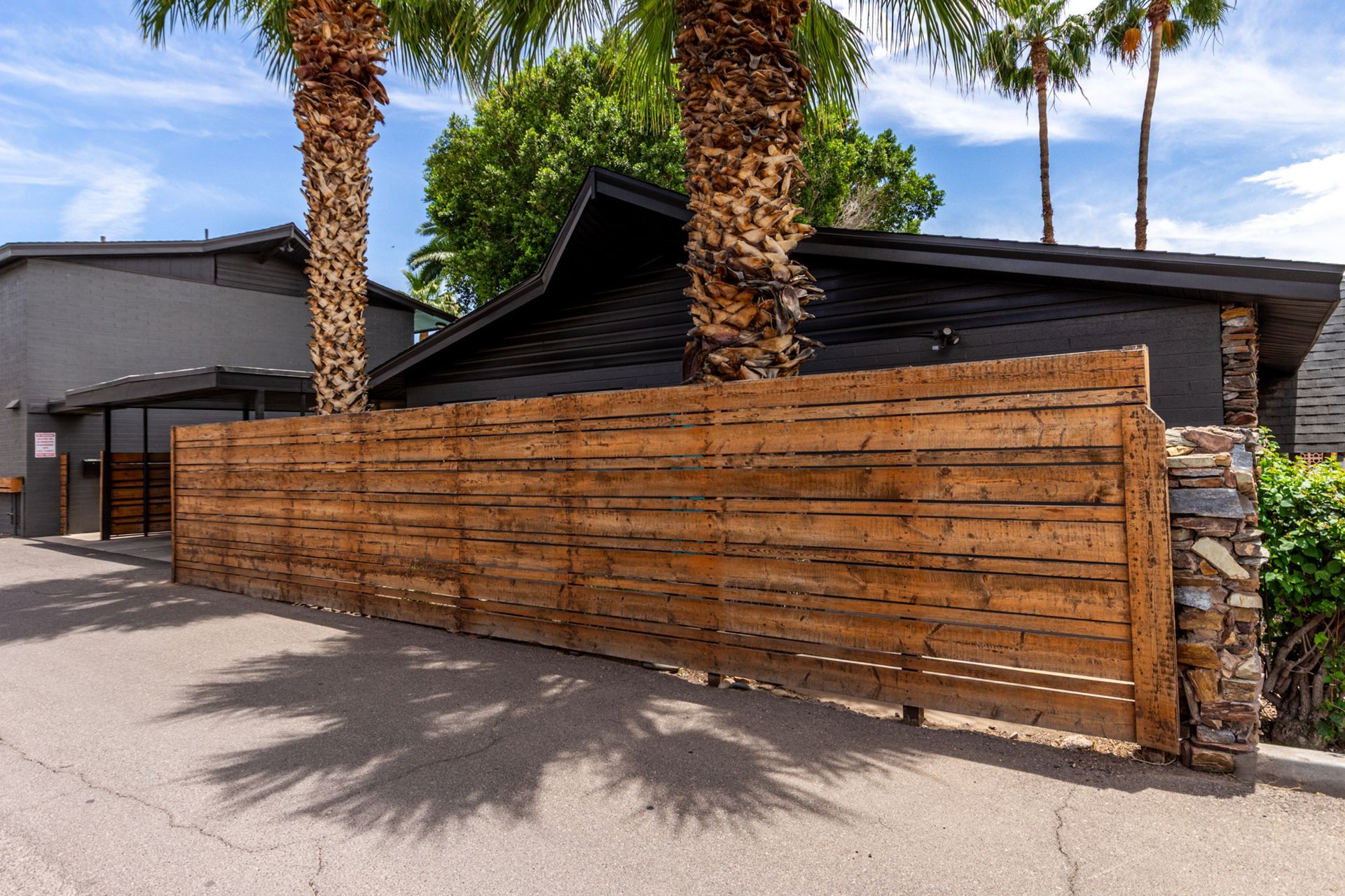 Wooden fence with palm trees and a dark-colored building behind it on a sunny day.