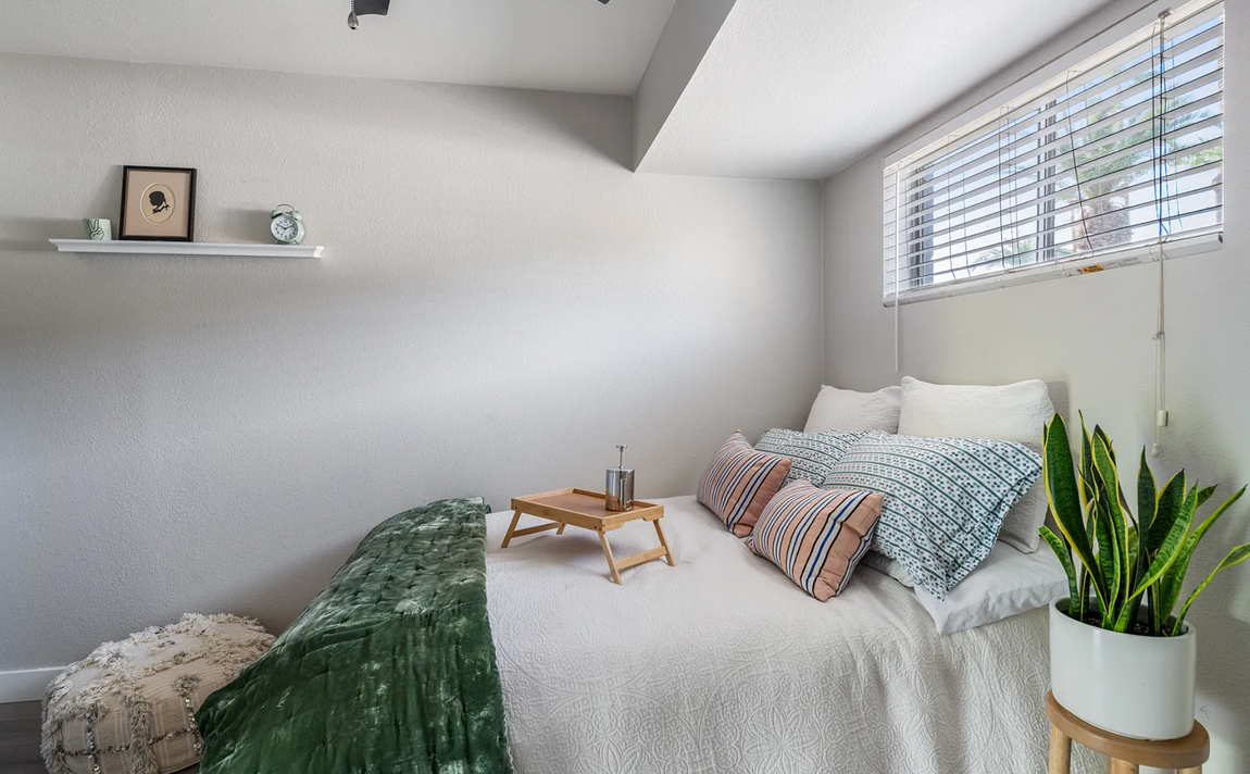 Bedroom with bed, pillows, blanket, tray with drinks, small shelf with decor, and a potted plant.