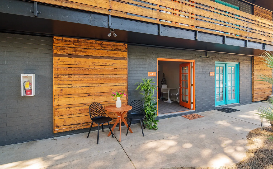 Exterior of a building with wooden and grey walls, orange and turquoise doors, and outdoor seating.