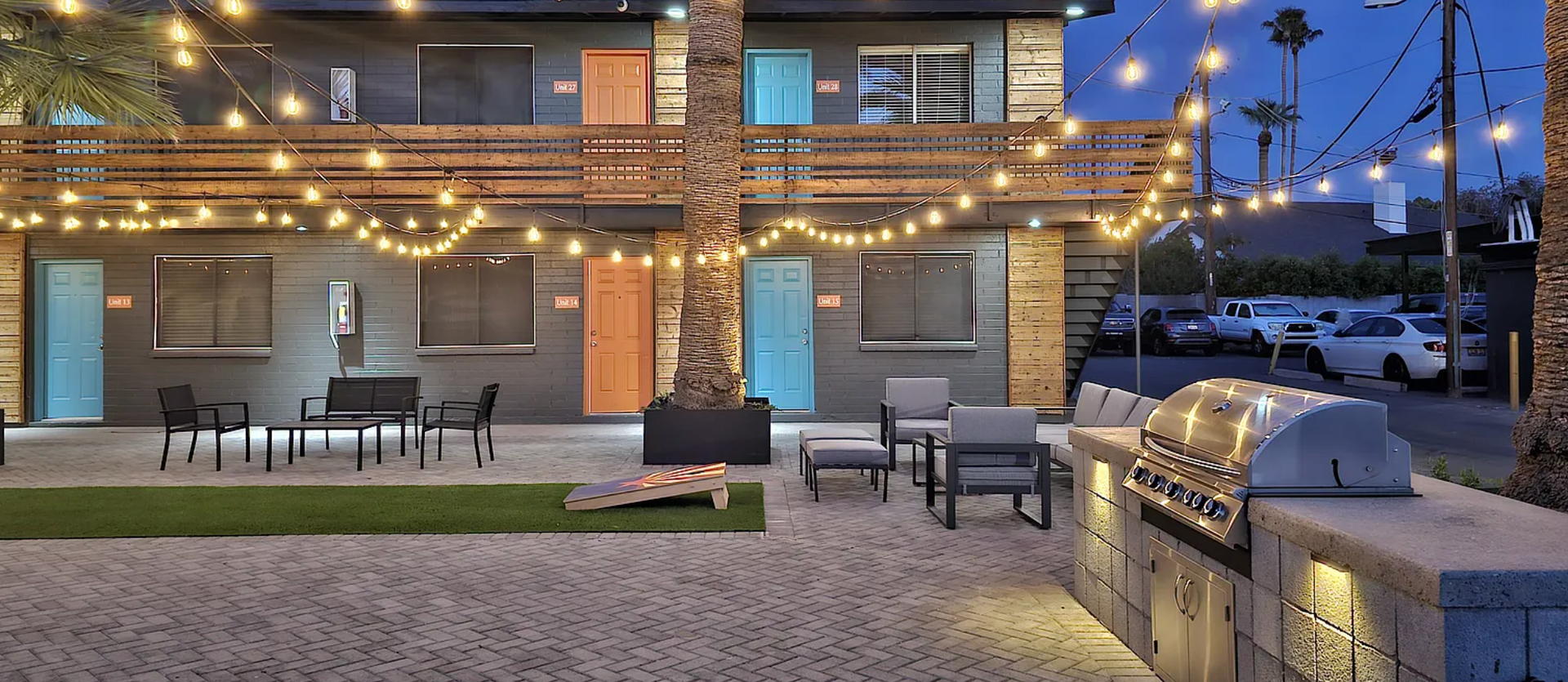 Apartment courtyard with string lights. Grey building with colorful doors, outdoor seating, and grill. Evening setting.