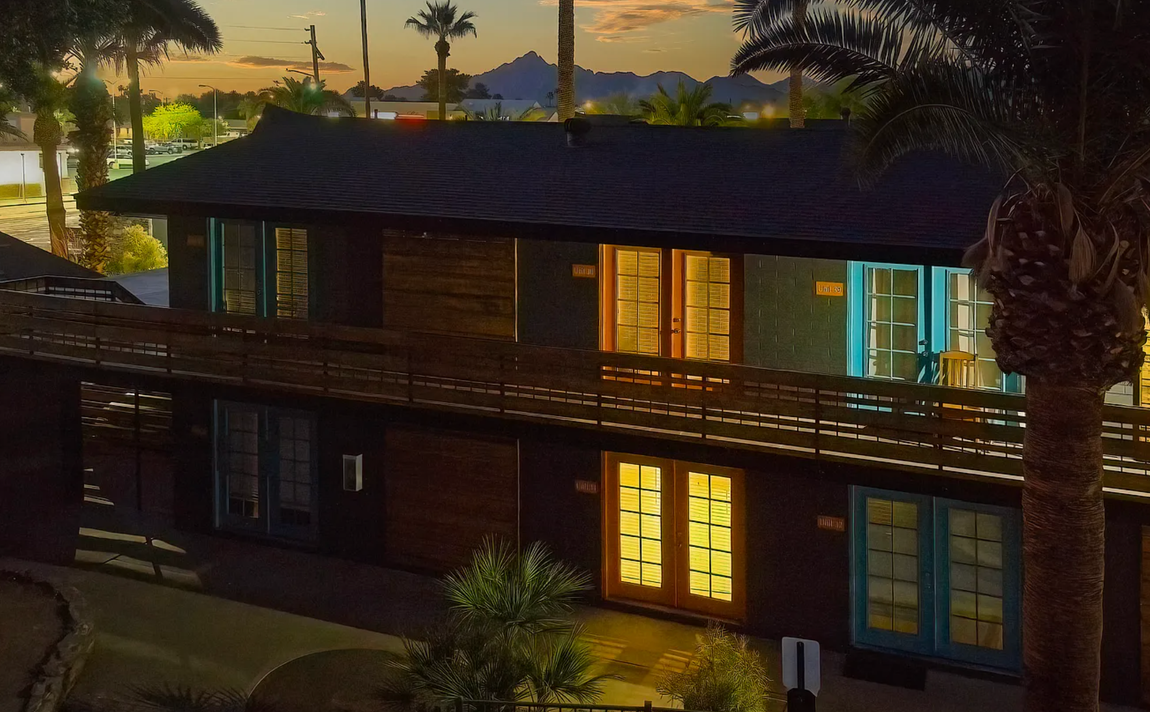 Two-story dark building at dusk with lit windows, palm trees, and mountain backdrop.