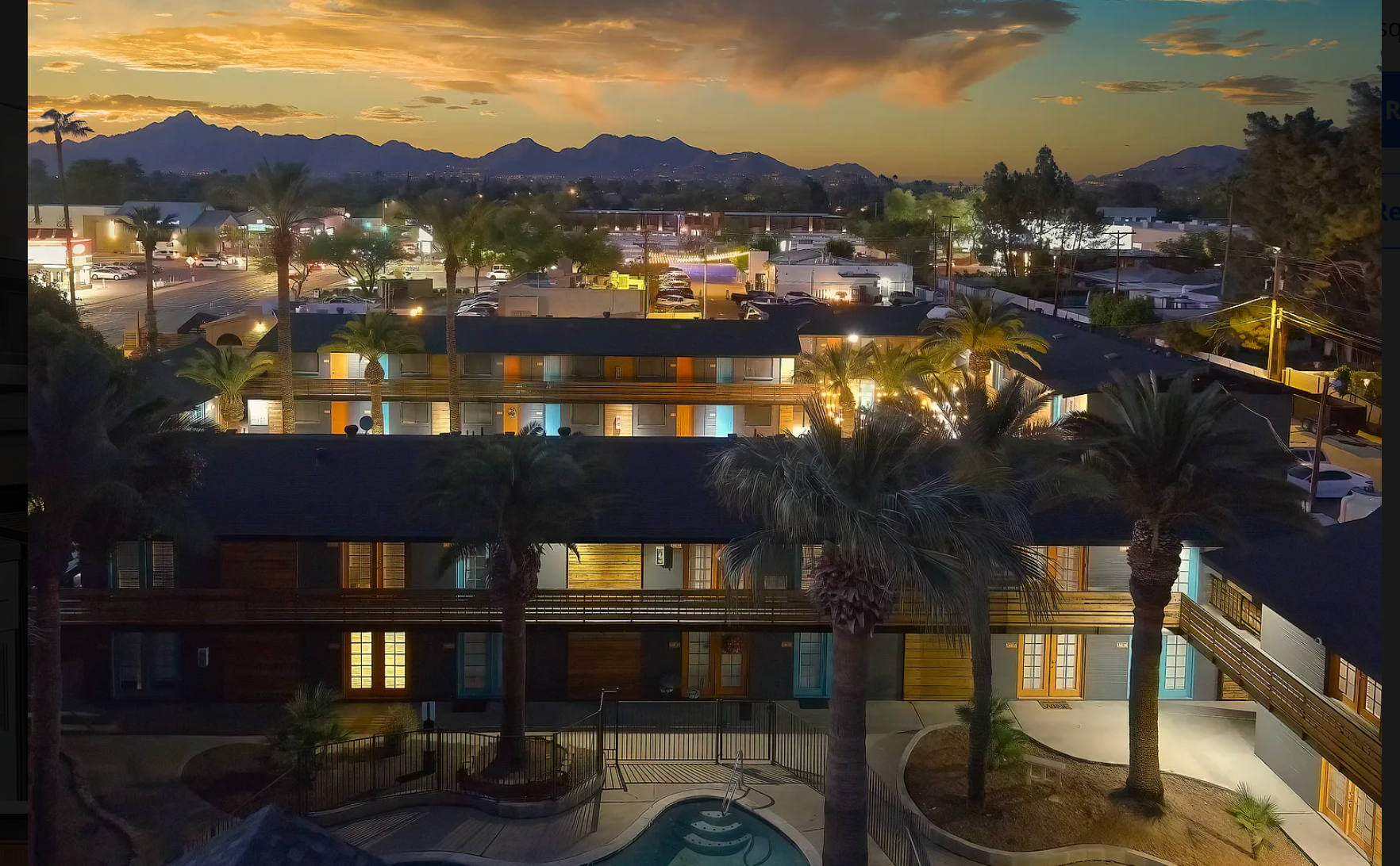 Aerial view of a hotel at dusk, palm trees, mountains, town lights, and a pool.
