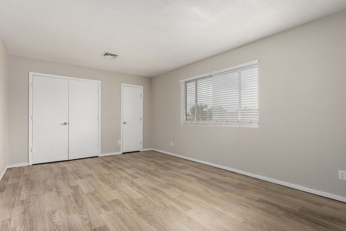 Empty bedroom with light wood-look flooring, two white closet doors, a small door, and a window with blinds.