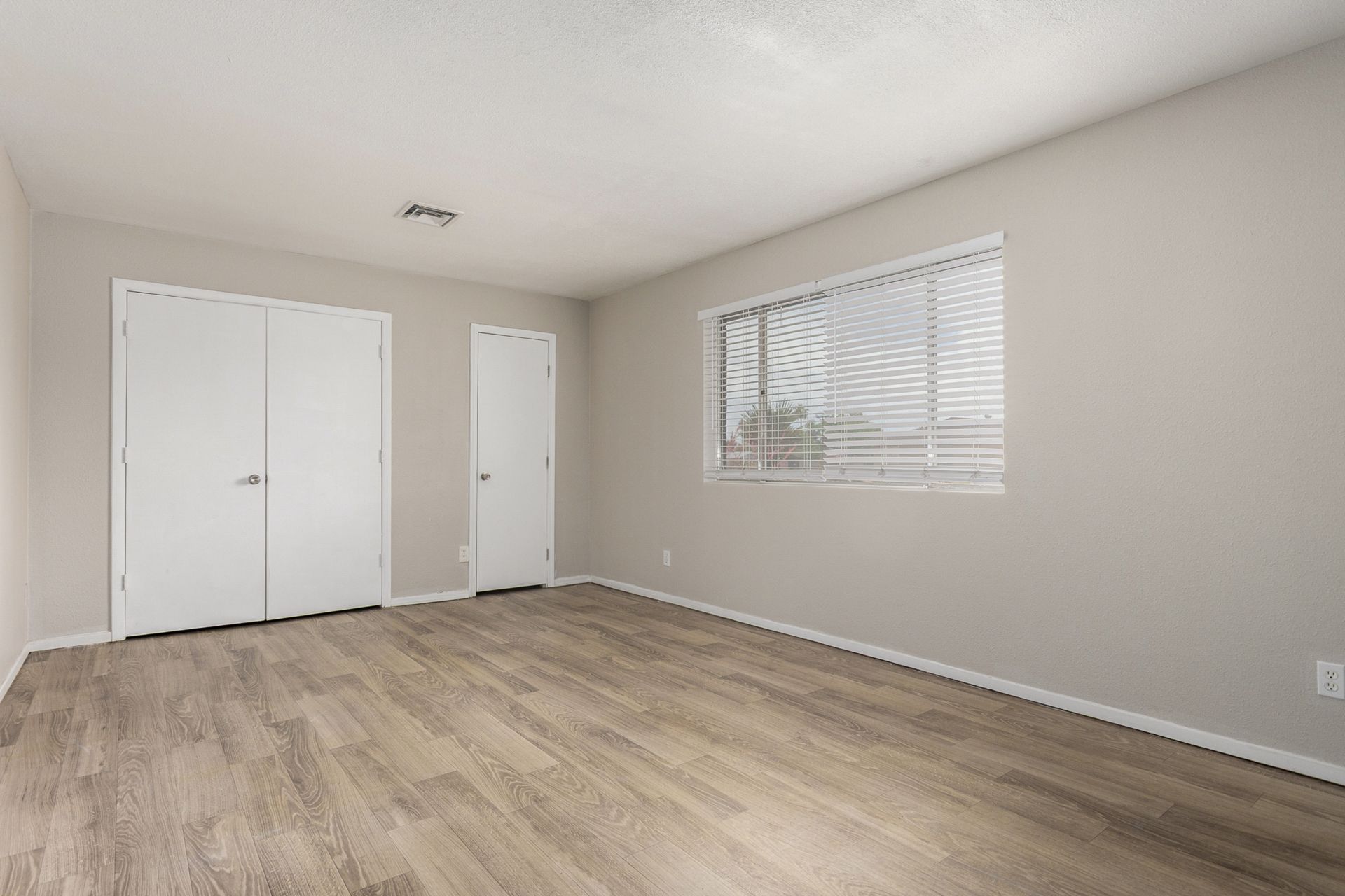 Empty bedroom with light wood-look flooring, two white closet doors, a small door, and a window with blinds.