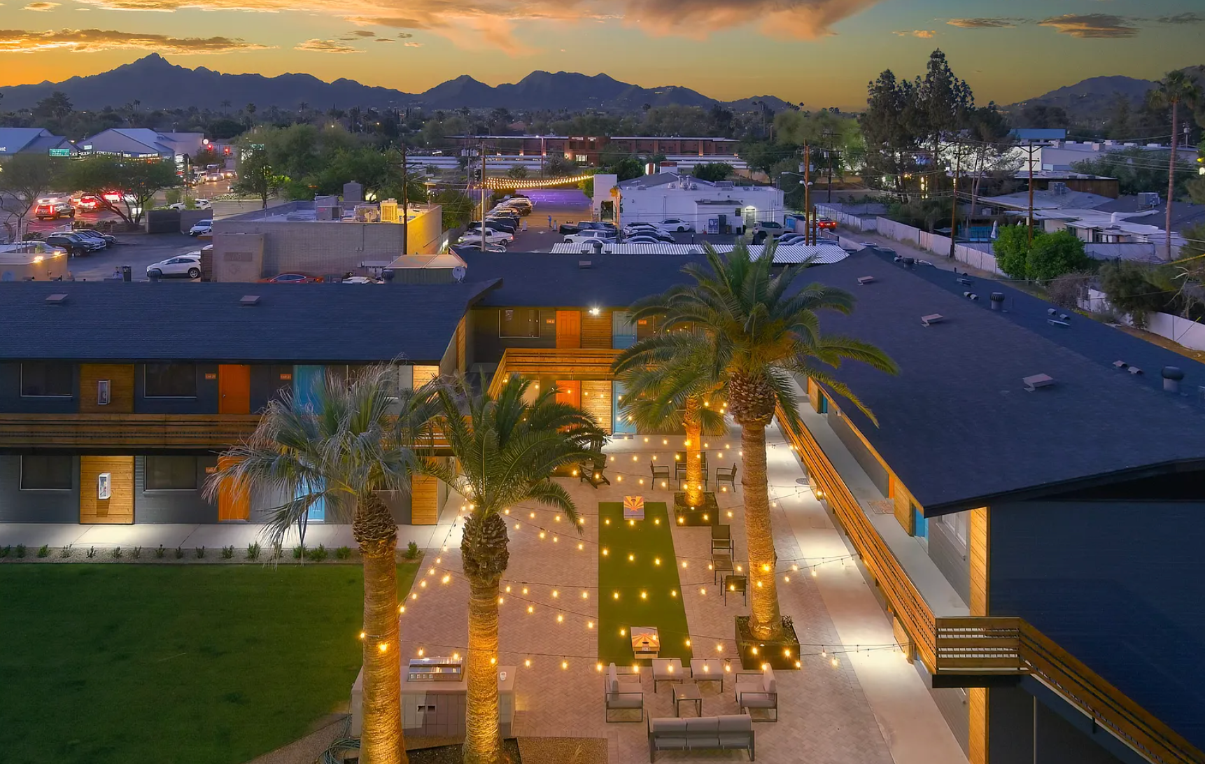 Aerial view of a brightly lit courtyard with palm trees, tables, and a two-story building at sunset.