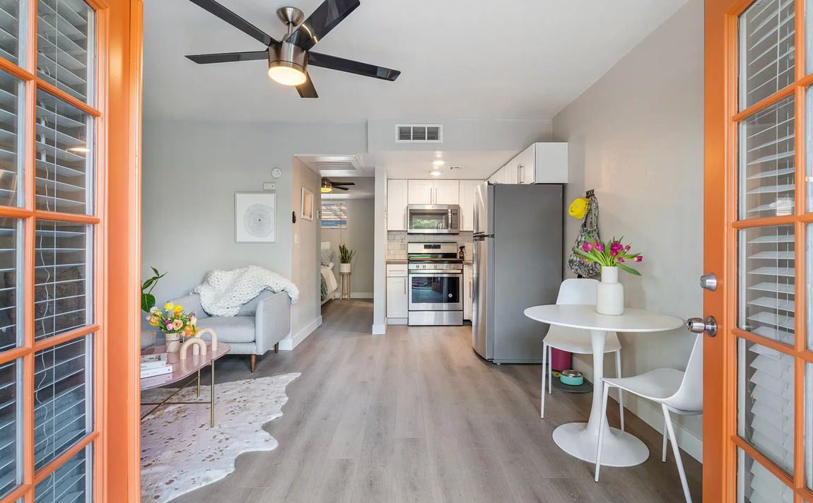 Small apartment interior with kitchen, dining area, and sofa. Orange door frames, gray walls, wood floors.