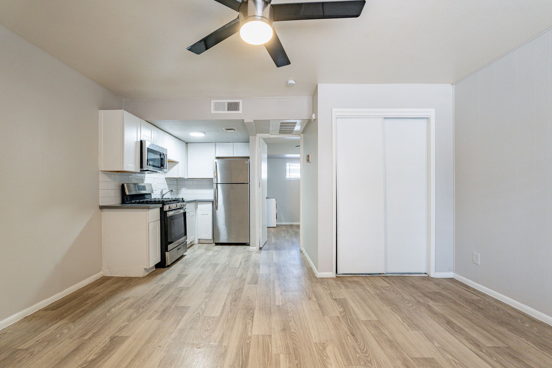 Empty apartment interior with kitchen, closets, and ceiling fan. Light wood flooring and neutral walls.