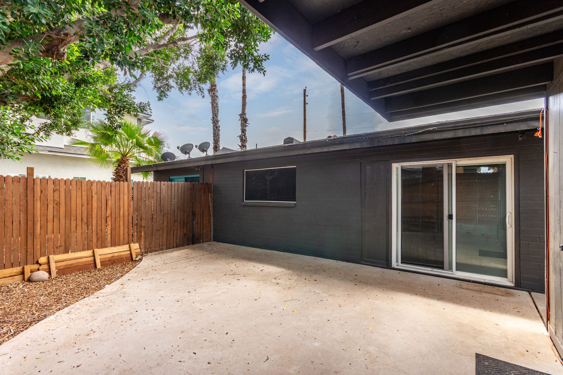 Backyard patio with gray building, sliding glass door, wooden fence, trees, and blue sky.
