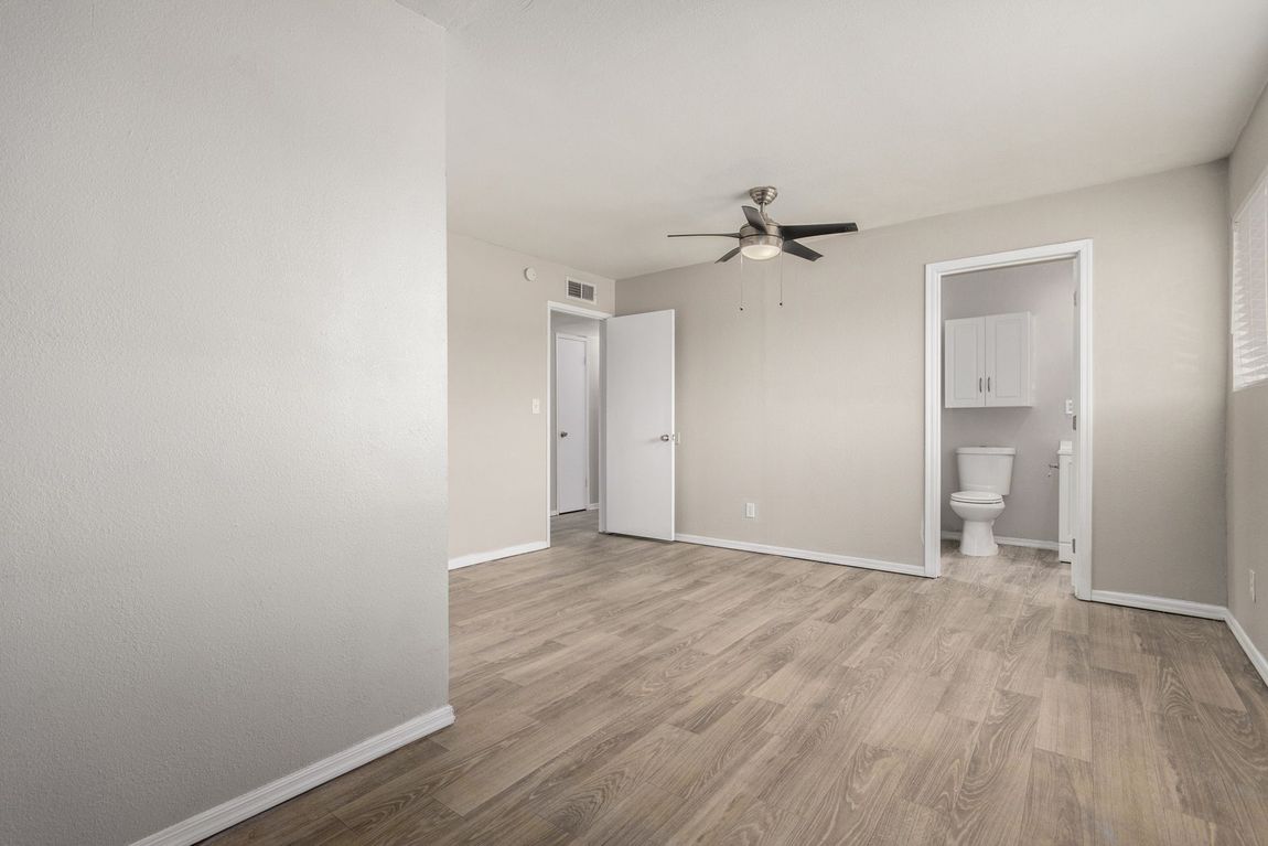 Empty bedroom with wood-look floor, doorways, and a bathroom. Neutral walls and a ceiling fan.
