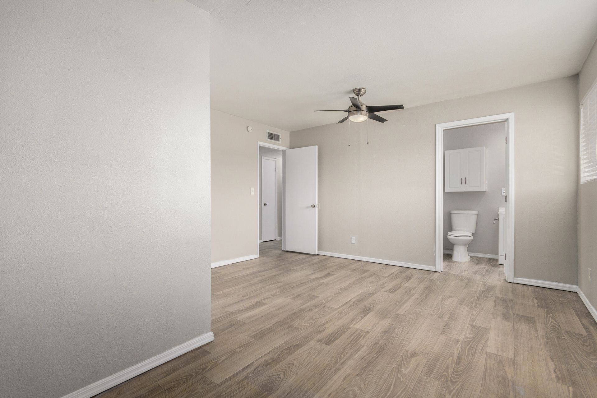 Empty bedroom with wood-look floor, doorways, and a bathroom. Neutral walls and a ceiling fan.