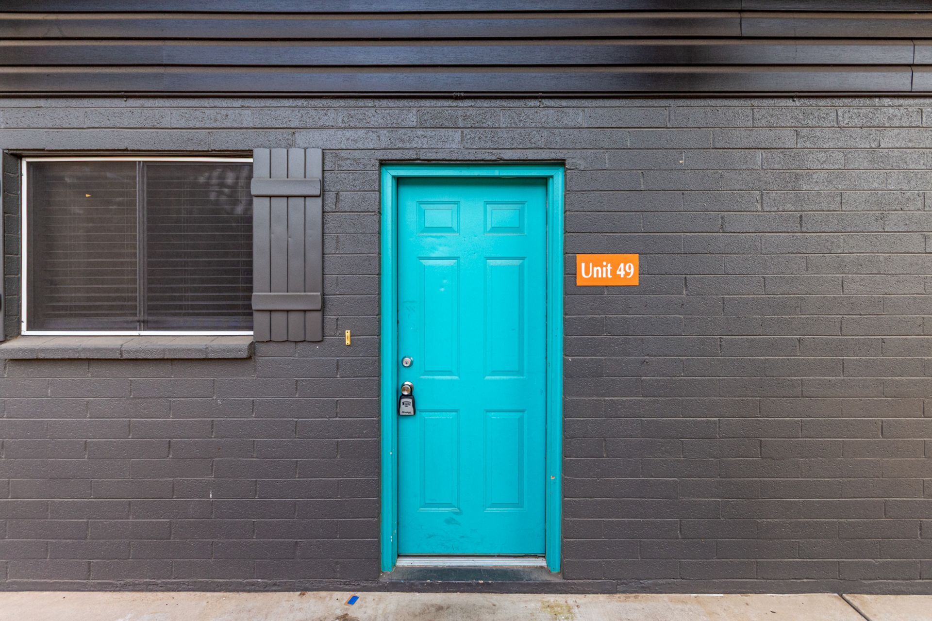 Black brick building with a bright teal door, window with shutter, and orange sign.