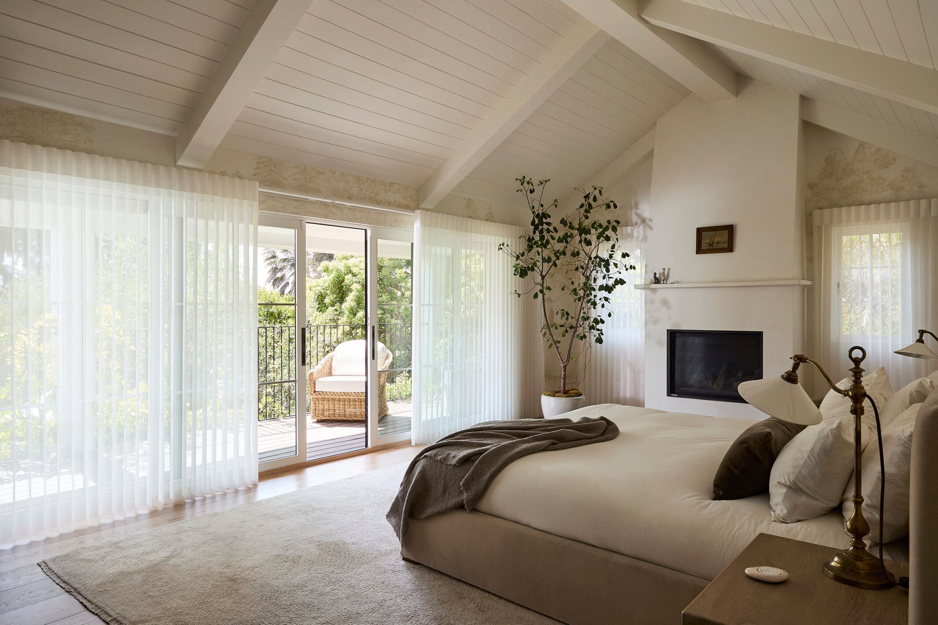 Bright bedroom with vaulted ceiling, large windows, white curtains, beige bed, and fireplace.
