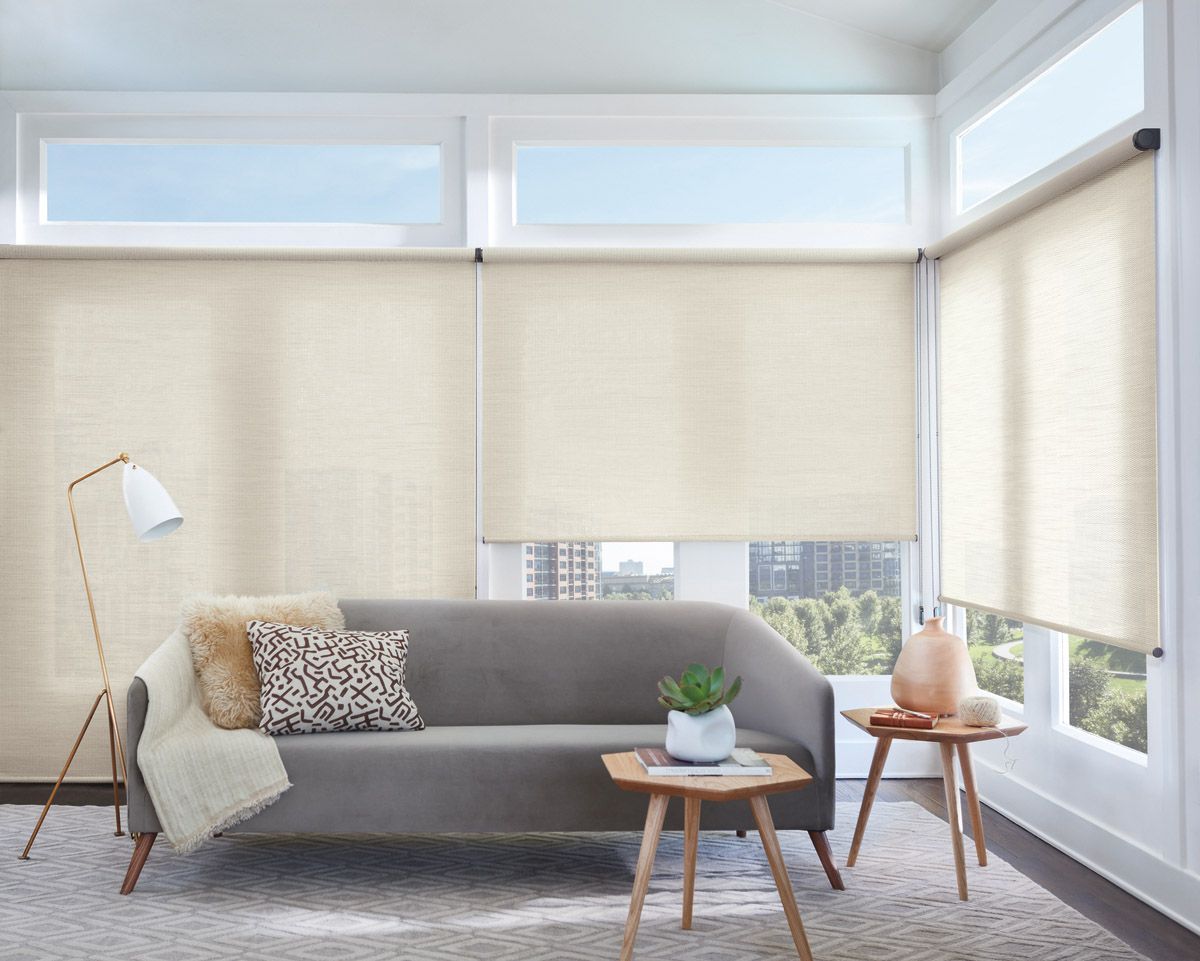 Bright living room with beige blinds, gray sofa, floor lamp, and small wooden tables by large windows
