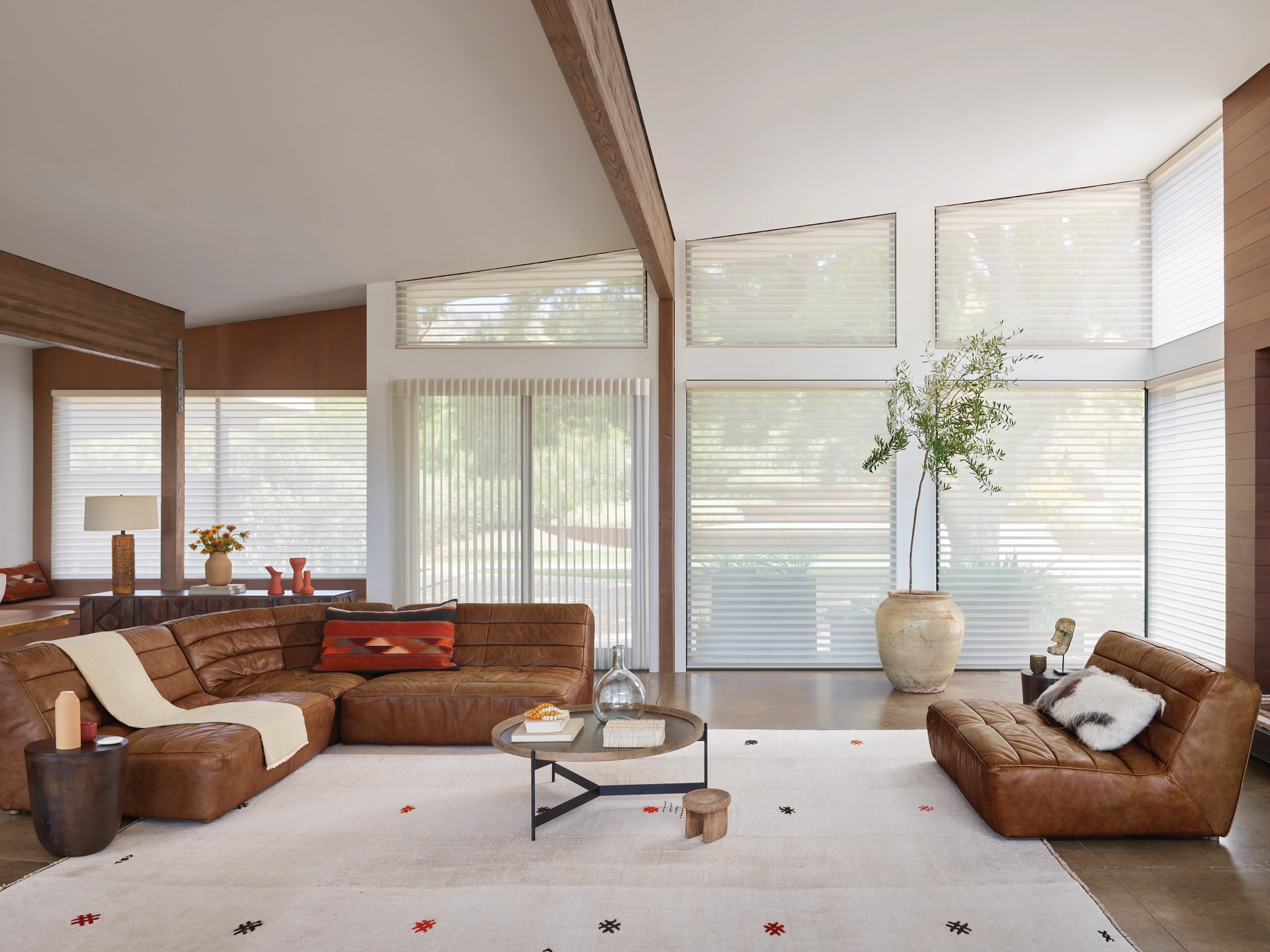 Bright living room with large windows, brown sofas, white rug, and a tall vase of flowers