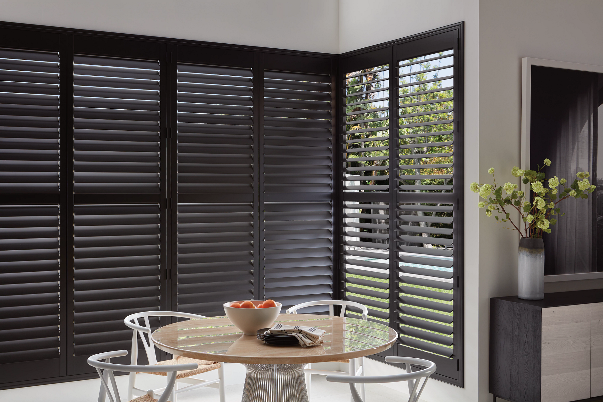 Dining area with round table and chairs beside dark louvered blinds, with a vase on a side console.
