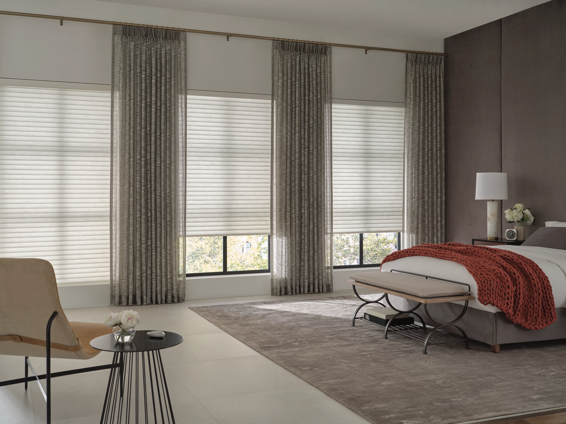 Modern bedroom with large windows, gray curtains, beige chair, white bed, and red throw blanket