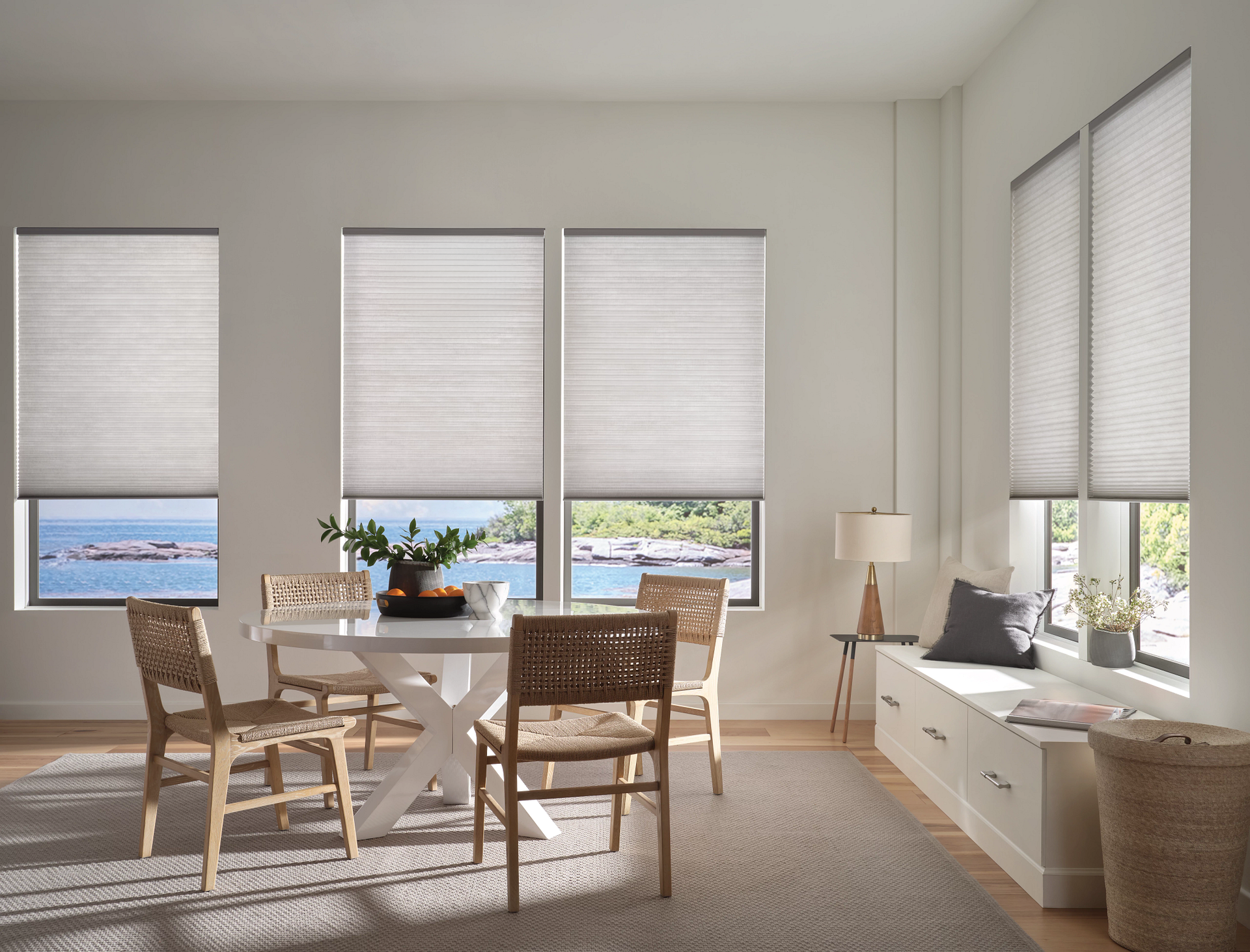 Bright dining nook with wicker chairs, white blinds, and a bench by sunlit windows overlooking water.