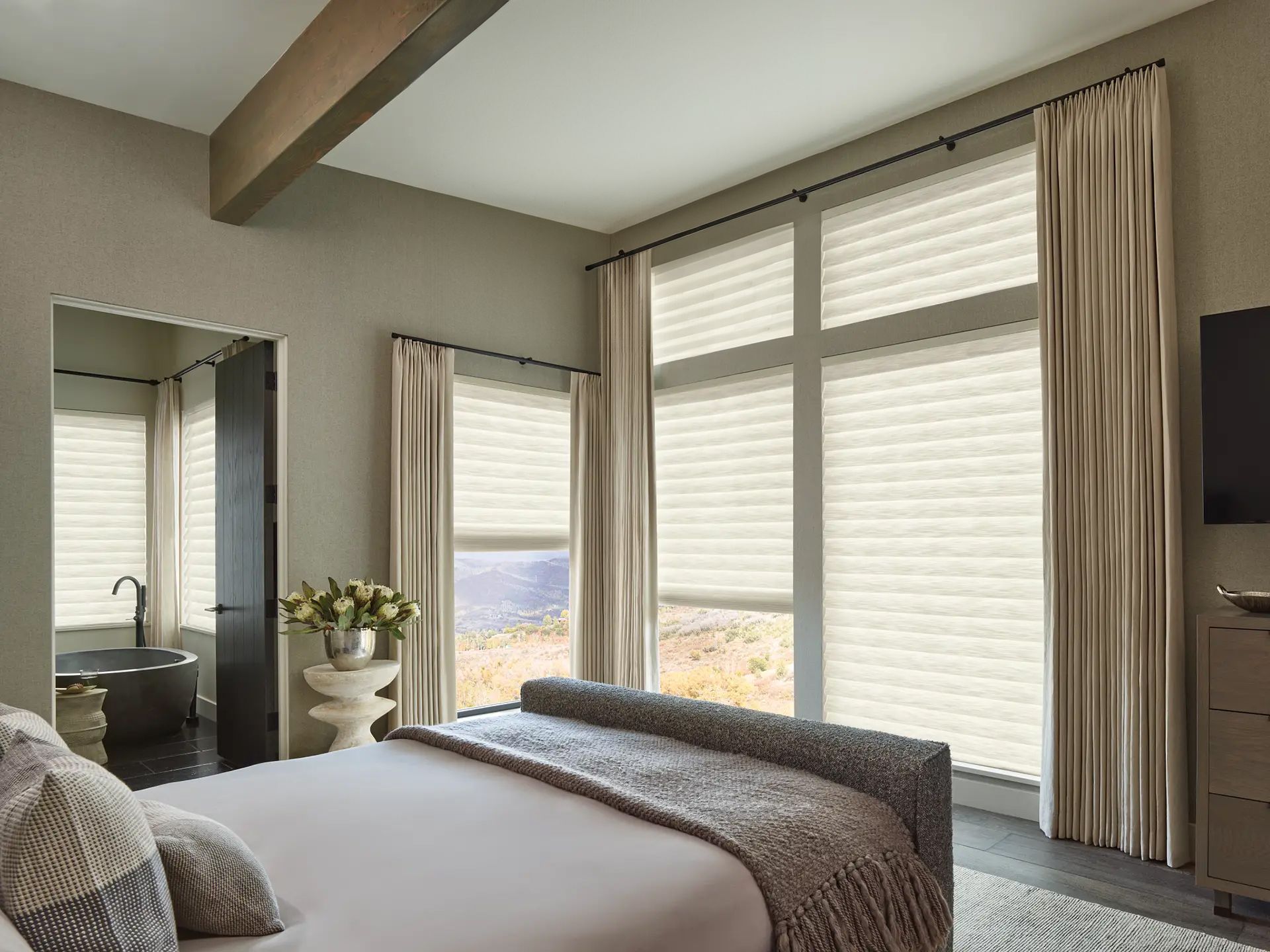 Modern bedroom with large windows, beige bedding, mirrored dresser, and sheer curtains overlooking a view outside