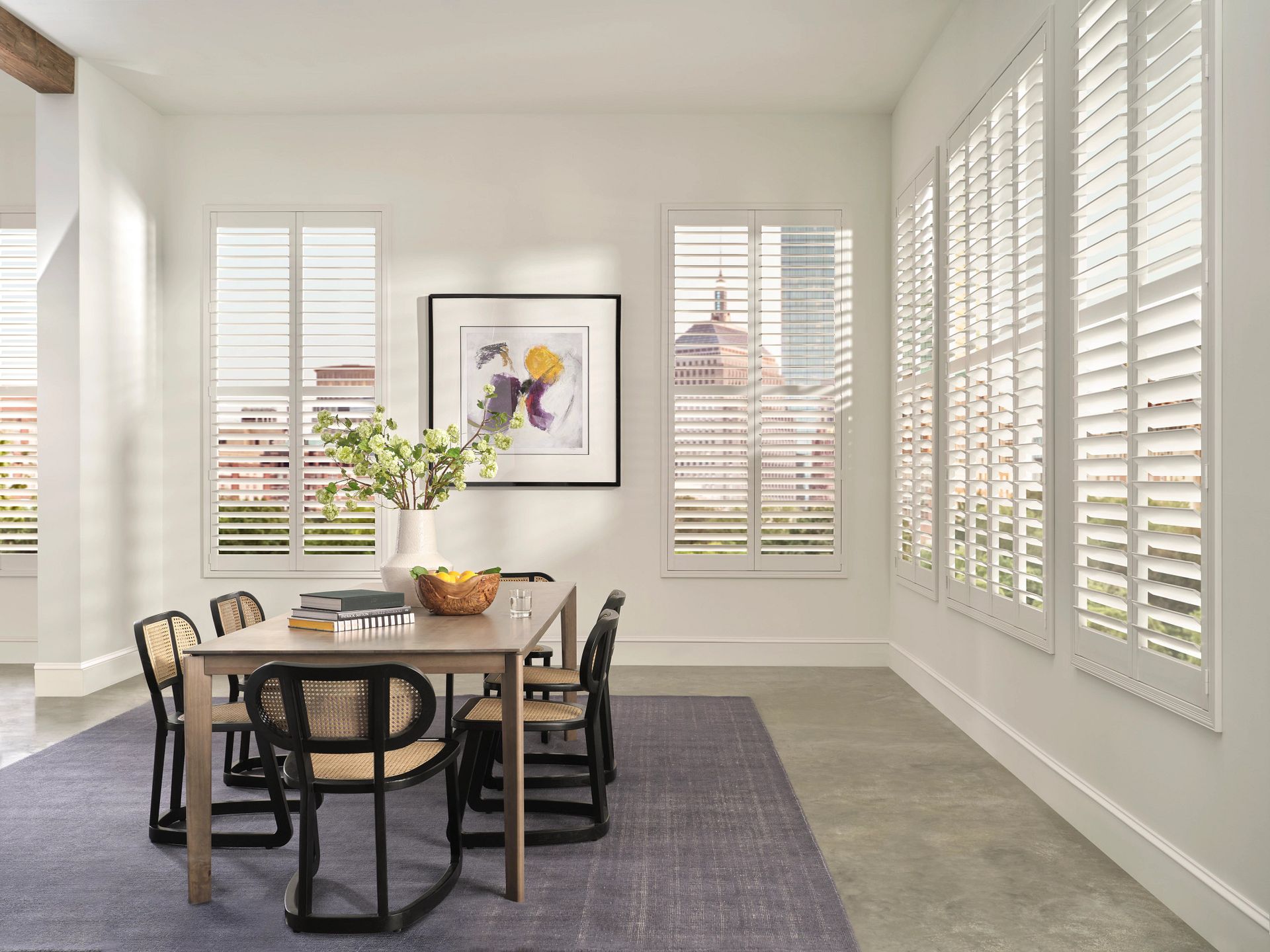 Bright dining room with wooden table, black chairs, large windows, and sunlight on a gray rug.