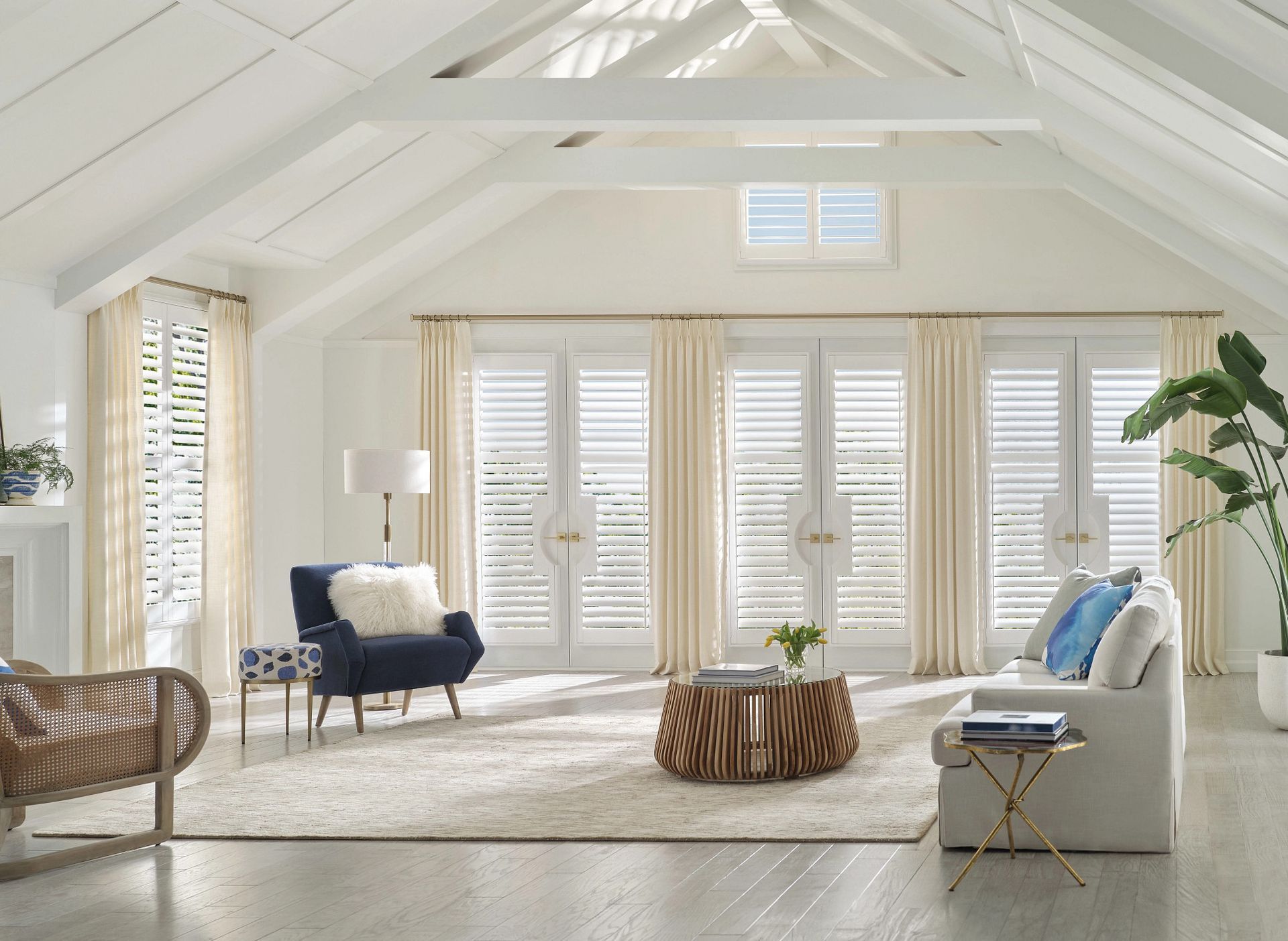 Bright white living room with vaulted ceiling, shuttered windows, blue chairs, and a rustic wood coffee table