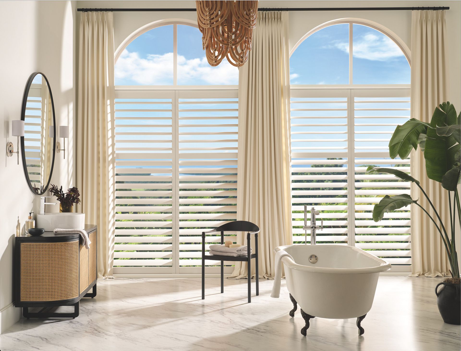 Bright sunlit bathroom with arched windows, a freestanding tub, vanity, and cream curtains