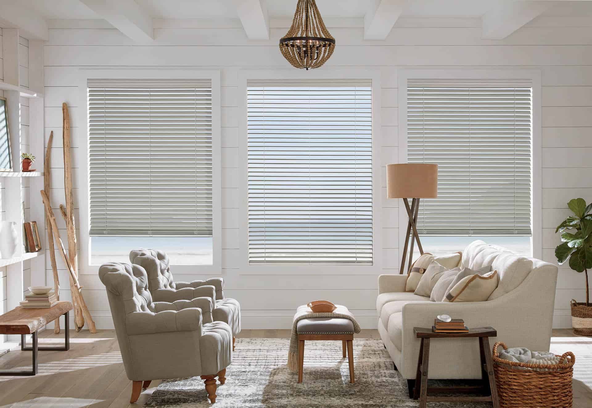 Bright living room with white walls, three large windows, beige armchairs, sofa, rug, lamp, and wicker basket.