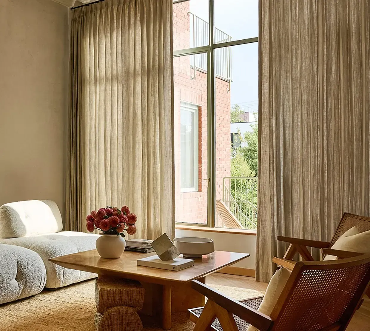 Sunlit living room with beige curtains, wooden chairs, sofa, and a coffee table with flowers