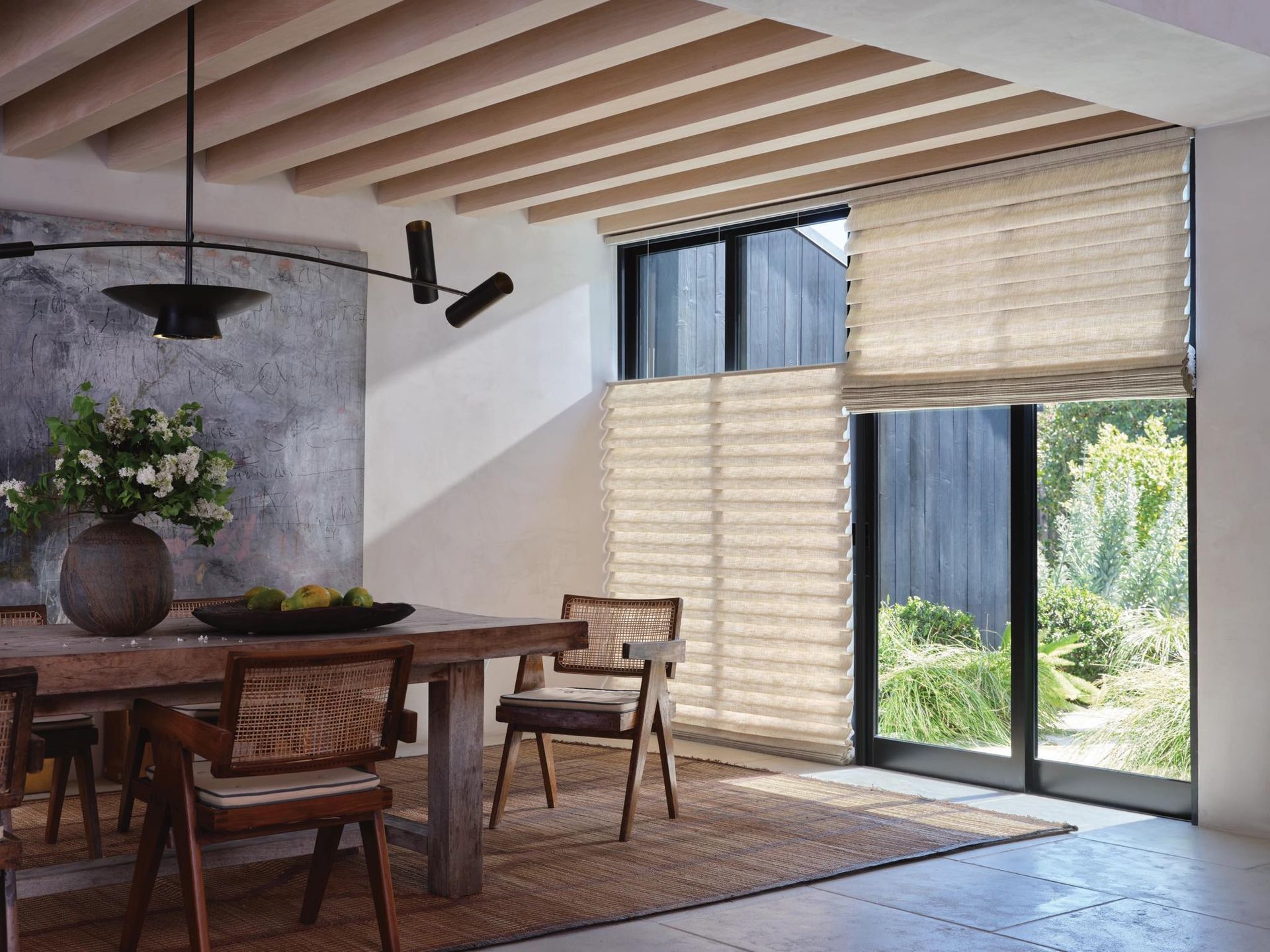 Modern dining room with wooden table, woven chairs, exposed ceiling beams, and large windows with blinds.