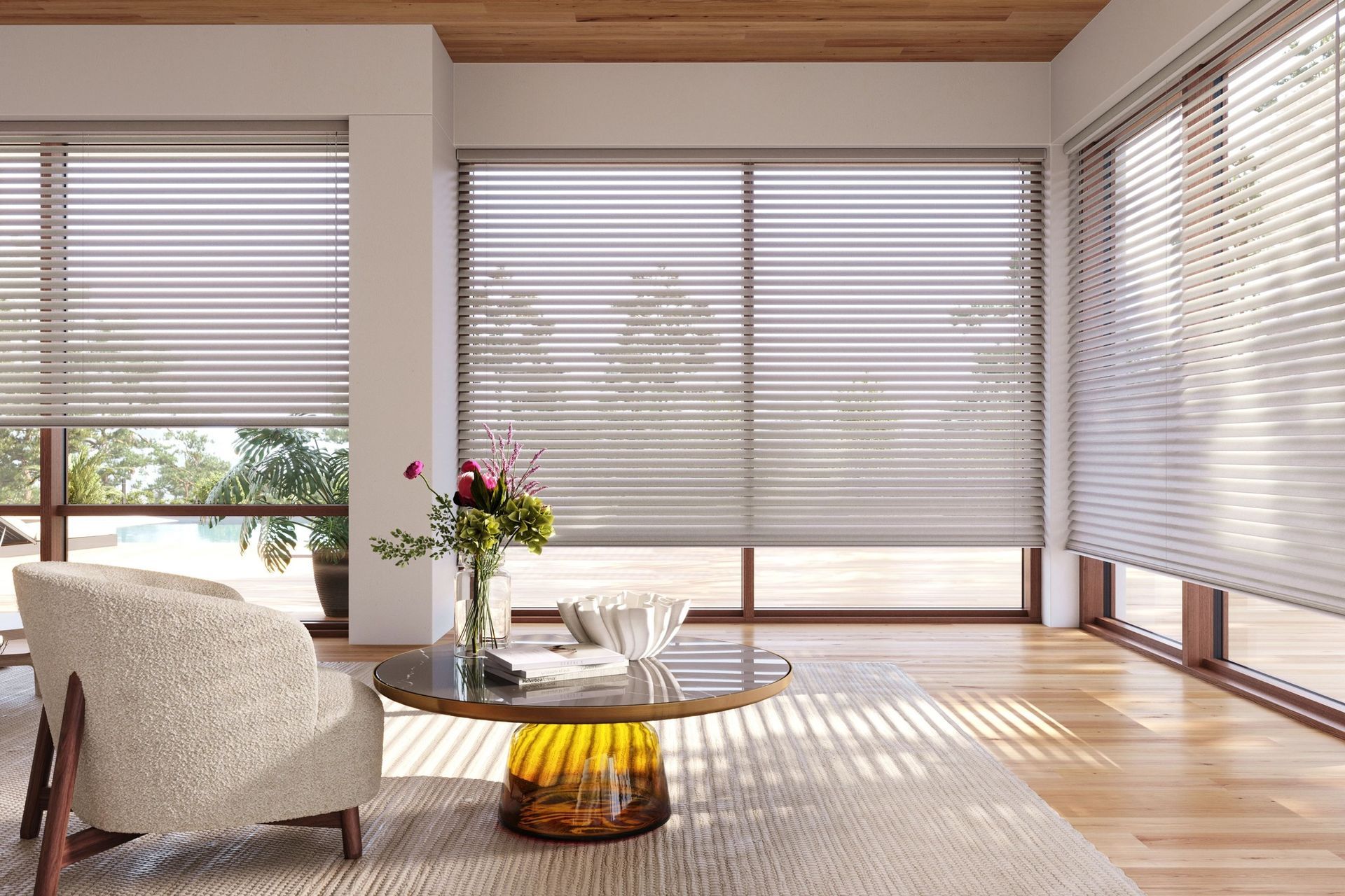 Bright sunlit living room with large windows, blinds, a white chair, rug, and vase of flowers on a table